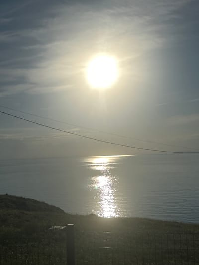 The image depicts a serene seascape under a bright, hazy sun. The sun, positioned in the upper center, casts a strong glare and its light reflects intensely on the water, creating a shimmering path that stretches towards the horizon. The sky is a pale blue, with wispy clouds partially obscuring the sun. Two thin wires stretch diagonally across the upper portion of the frame. The foreground features a grassy slope, with a dark fence and a wooden post in the lower right quadrant. The overall impression is one of a tranquil, sunlit coastal landscape. The specific location of Мысовское сельское поселение, Ukraine, is not visually discernible from the image content.