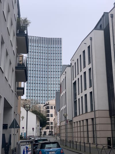 The image shows a narrow street in Frankfurt am Main, Germany, with a tall office building in the background. The street is lined with residential buildings on both sides. The buildings are mostly gray and white with a few black accents. The street is paved with asphalt and has a few parked cars. The streetlight posts have a black base and a silver lampshade. The street is empty except for a blue Mini Cooper that is parked in the street. The office building is 10 floors and has large glass windows. The buildings in the foreground have some plants growing on the balconies and in the ground. The weather is overcast.