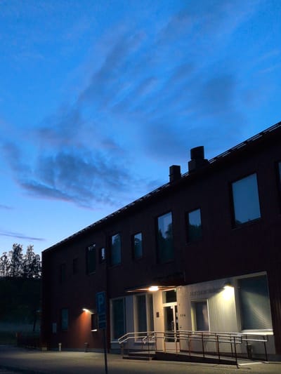 The image depicts a dark, modern building, possibly an office or public institution, with a dark red-brown exterior and numerous rectangular windows. The building has a flat roof with two dark chimneys and a dark railing along the roofline. The sky above is a deep, twilight blue with wispy, dark clouds. To the left of the building, a line of silhouetted trees is visible against a dark, hilly background.

In the foreground, there is a paved area, likely a sidewalk or road, with a lamppost on the left. A blue parking sign with a white "P" is visible on the right side of the building. The building features a prominent entrance area with a ramp and a railing, illuminated by overhead lights. The facade of this entrance area is clad in light-colored, ribbed siding. Some windows have blinds. The number "00A" is visible near the entrance.

The overall impression is one of a commercial or institutional building in a semi-rural or suburban setting at dusk. Based on the architectural style and potential signage, the building is likely located in Vantaa, Finland.