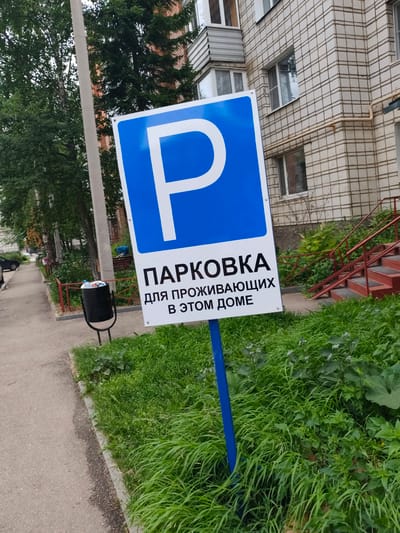 The image shows a blue and white parking sign in a residential area. The sign has a large white "P" on a blue background, followed by text in Russian that reads "ПАРКОВКА ДЛЯ ПРОЖИВАЮЩИХ В ЭТОМ ДОМЕ," which translates to "PARKING FOR RESIDENTS OF THIS BUILDING." The sign is situated on a blue metal pole, in front of a patch of green grass and vegetation. In the background, a multi-story building with light-colored brickwork and several windows is visible. To the left of the sign, a sidewalk runs alongside a street with trees and a black trash can filled with refuse. A car is partially visible on the street. The overall scene suggests a typical urban environment in Russia, likely a residential courtyard. The specific location of Syktyvkar, Russia, is not explicitly indicated by any visual cues in the image itself, but the context of the Cyrillic alphabet on the sign strongly suggests a Russian-speaking country.