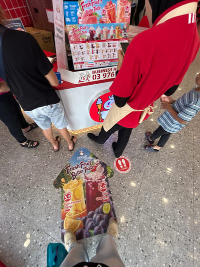 The image captures a top-down view of people queuing or standing near a Mixue drink stall in an indoor commercial setting, likely within a shopping mall in Subang Jaya City Council, Malaysia. The floor is tiled with a speckled grey pattern.
Subjects include several individuals, mostly visible from the waist down. An adult in a red jacket and black pants stands close to the counter, accompanied by a child in a striped shirt and dark pants. Other patrons include a person in light blue shorts and flip-flops, another in black pants and flip-flops, and an individual in light blue pants and white shoes, standing further back next to a teal shopping bag.
The primary activity is waiting to order or receive beverages from the Mixue stall. This is indicated by the positioning of people and a prominent red circular floor sticker that reads "PLEASE STAND HERE" with white footprints, suggesting a designated queuing spot for social distancing.
Notable details include the vibrant advertisements for Mixue's products, especially the "Fresh Fruit Better Taste" series. Menu items like "Strawberry Bobo," "Peach Bobo," "Fresh Punched Orange," and "Taro Grape Tea" are displayed with prices in Malaysian Ringgit (RM). Categories such as "Ice Cream Series," "Fruit Tea Series," "Milk Tea Series," "Coffee Series," and "Pure Tea Series" are also visible. Text also includes details about product handling like "Frozen After Harvest," "-35°C Frozen," and "-18°C Cold Chain Transport," alongside a "No Customization Product Allowed" sign. A business contact or identification "BUSINESS H 03 976" and an "Alipay+" logo are present. Chinese text for "Snow King Crispy Fries (Honey Butter Flavor)" also appears on one of the signs.