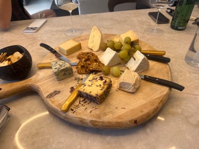 An overhead shot of a cheese board on a marble table. The cheese board is a light wood and is oval shaped. On the board are several types of cheese, grapes, and crackers. There are also two knives with black handles and two knives with gold handles. A dark bowl with crackers is to the left of the board. In the background, two glasses of white wine, a Camelbak water bottle, a phone, and a set of keys are visible. The overall setting appears to be a restaurant. The image is taken from a high angle, looking down at the table. There is no indication of the location being Ridderkerk, Netherlands.
