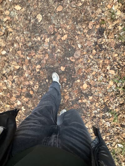 A person wearing gray sneakers and black jeans stands on a ground covered in fallen leaves. The leaves are mostly brown and tan, scattered across a dirt path. Part of a black jacket is visible on the right side of the frame. The image is taken from a slightly elevated, downward-facing perspective, offering a view of the ground and the person's lower body.