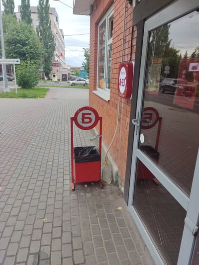 An outdoor scene in Serpukhov, Russia, features a sidewalk and the exterior of a brick building with large glass doors and windows. In the foreground, a red trash receptacle with a black liner stands on a paved sidewalk made of interlocking gray bricks. A red metal frame surrounds a circular sign with a white background and a large Cyrillic letter "Б" in red. To the right of the trash can, a plastic bottle filled with liquid is partially visible. The building's facade is constructed of red brick, and a round red sign with white lettering, indicating business hours (8-23), is mounted on the wall. Reflections in the glass doors show trees and the street. In the background, a multi-story building and more trees are visible. A street sign indicating "ул. ВОРОШИЛОВА" (Voroshilova Street) is also present. The sky is overcast.