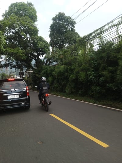 A car and a motorbike travel on a paved road. The car, a black van with a license plate reading "F 1021 GU", is directly to the left. A person on a motorbike is directly ahead of the car in the lane. The person is wearing a dark jacket, backpack, and a helmet. A solid yellow line is present on the road, near the center of the road. Lush trees and greenery are to the right, and more trees are in the background. Electrical wires are visible in the sky. The location might be in Taunggyi, Myanmar.