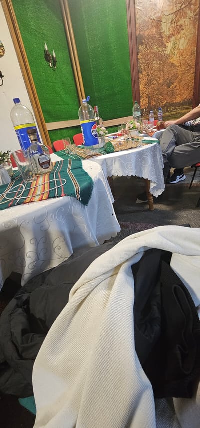 The image shows a table setting in what appears to be a restaurant. Two tables covered in white patterned tablecloths are visible, with a person seated at the far table. One table is partially covered with a green, red, and yellow striped runner. Empty plastic bottles of Pepsi and other drinks are placed along both tables, with glasses scattered between them. The background features a framed painting of a forest and a section of artificial grass resembling a wall decoration. Other items include a white and black garment pile. Given the query context, the location is presumed to be in Sveti Konstantin, Bulgaria.