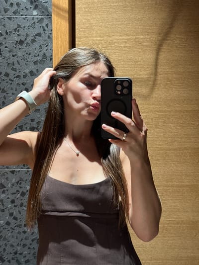 A young woman with long, straight brown hair is taking a selfie in a bathroom. She is wearing a dark brown tank top and a light green smartwatch. She is holding her phone in her right hand and her left hand is in her hair. She is wearing a silver necklace with a small pendant and a gold ring on her left ring finger. The background is a textured grey tile wall and a wooden partition. The lighting suggests it is daytime, with sunlight casting shadows on the wall.