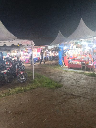 Under a dark night sky, a bustling outdoor market is illuminated by bright lights under white tents. Merchandise is displayed on tables and racks, and a person walks through the center aisle. Several parked motorcycles are visible in the foreground on the left side of the image. The ground is muddy.