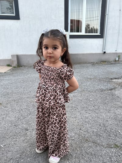 In Slanchevo, Bulgaria, a young girl with dark hair tied into two ponytails with white bows stands on a gravelly surface in front of a white building. She is wearing a pink leopard-print outfit consisting of a smocked top with puffed sleeves and wide-leg pants. Her hands are clasped behind her back, and she is looking directly at the camera with a neutral expression. The building behind her has a dark-trimmed window and a downspout. The environment appears to be an outdoor, possibly residential, area.