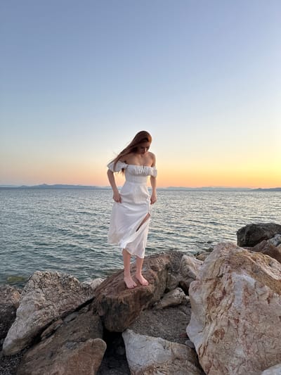 A young woman with long, auburn hair stands on large rocks by the sea. She is wearing an off-the-shoulder white dress with a slit up the side, which is blowing in the wind. Her bare feet are visible, and she is looking down at her dress. The sea is calm, with small waves rippling towards the shore. In the background, a hazy sunset paints the sky in hues of orange, pink, and yellow, with distant mountains visible on the horizon. The foreground is dominated by a rocky shoreline. The scene appears to be captured at sunset, with the sky showing the transition from day to evening. This location is likely Paleo Faliro, Greece, known for its coastline and views.