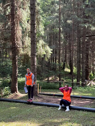 In a sun-dappled forest, two individuals are engaged in an outdoor activity. One person, wearing an orange vest over a pink jacket and dark pants, is suspended horizontally by a rope, their legs extended. They are also wearing white boots and sunglasses. Nearby, another person in a similar orange vest over a grey jacket, dark pants, and pink shoes, stands holding a clipboard. They appear to be observing or participating in the activity. The setting is a dense forest with tall trees and lush green undergrowth. Two thick, dark, cylindrical objects are visible on the ground, possibly part of an obstacle course or equipment for the activity. The presence of the vests suggests a structured event or team activity. The lighting indicates it is daytime, with sunlight filtering through the trees, casting shadows. The environment appears natural and somewhat rugged, characteristic of a forest location.