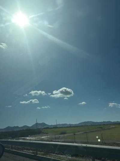 The image displays a sunny daytime landscape in the vicinity of Nzalat Laadam, Morocco, observed from inside a vehicle. The upper two-thirds of the frame features a clear blue sky with scattered white cumulus clouds. Intense sunlight originating from the upper left quadrant produces a prominent sunburst and lens flare, alongside reflections visible on what appears to be a vehicle's windshield. The lower third of the image is occupied by a dark grey metal guardrail running horizontally, with a portion of a vehicle's side mirror or frame visible in the lower-left corner. Beyond the guardrail, extensive green fields extend into the mid-ground, partially bordered by a fence with visible posts. In the background, a range of distant mountains or hills is discernible, with two slender, tall structures, consistent with communication towers, positioned on one of the higher peaks. No individuals, animals, or explicit actions are depicted.