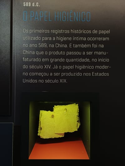 The image displays an information panel from an exhibit, likely in a museum, though the location is not specified in the image. The text, in Portuguese, provides a brief history of toilet paper. The exhibit mentions that the earliest recorded use of paper for personal hygiene was in 589 AD in China. The panel further notes that mass production of toilet paper began in China in the 14th century, and modern toilet paper production in the United States started in the 19th century. To the right of the text is a display case containing what appears to be a fragment of old, yellowed paper, possibly meant to represent early forms of toilet paper. The paper is presented against a black background with a section of orange visible behind the paper fragment.