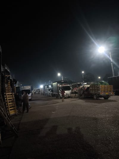 The image captures a nighttime scene in Pitalito, Colombia, depicting a busy street or market area illuminated by multiple bright streetlights. The environment consists of a dark sky above a paved roadway, with buildings or stalls lining the left side. Several vehicles, primarily cargo trucks of varying sizes, are present along the road, some appearing to be parked or in a queue.

On the far left, a stack of wooden pallets leans against a partially visible structure. Two adult males are observed in this area; one faces the street, while the other stands with his back partially to the viewer, facing the pallets.

In the mid-ground, a white cargo truck with a red accent on its cab is prominent, with a person in a red shirt walking in front of it towards the right. Further to the right, a reddish-brown open-bed pickup truck is parked, its bed filled with large green sacks, possibly produce. Several individuals are gathered around this truck, suggesting activity related to loading or unloading. Additional trucks are visible further down the street, extending into the background.

The powerful illumination from streetlights casts long shadows and produces significant lens flare from the brightest light source on the right side of the frame. The overall scene suggests commercial activity or transport operations occurring during late hours.