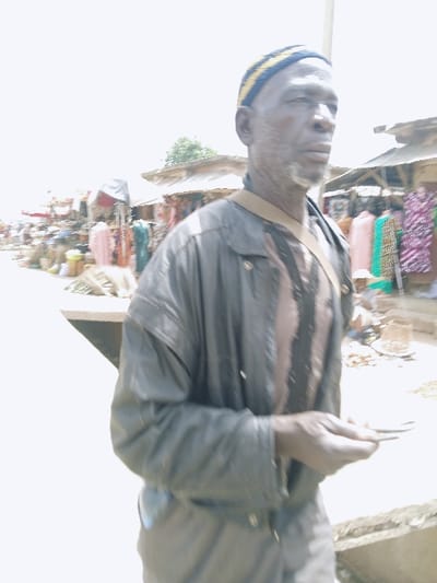 The image displays an outdoor market scene located in Kadoma, Zimbabwe, captured in bright daylight. An adult male with a dark complexion, appearing middle-aged to elderly, is the central figure in the foreground, positioned slightly to the right. He is wearing a blue and yellow striped kufi-style cap, a dark, long-sleeved jacket over a vertically striped garment, and a light-colored strap diagonally across his chest. He has a short, graying beard and mustache. His hands are visible, holding several small, dark, elongated objects. The man's gaze is directed towards his right, away from the camera. The background consists of a lively market area lined with multiple stalls featuring corrugated metal roofs, displaying various goods including vibrant textiles (pink, green, patterned fabrics) and containers. The ground appears uneven, likely dirt or earth, with a visible concrete edge in the lower left foreground. A utility pole is present behind the subject, and distant tree foliage can be observed above the market structures.