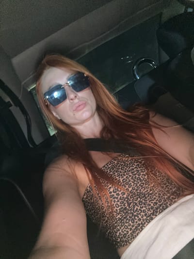 A woman with long, auburn hair is taking a selfie in the backseat of a vehicle, possibly on a road trip through Zagrađe, Montenegro. She is wearing oversized, angular sunglasses with a gold frame and a leopard-print tube top. Her lips are slightly parted, and she appears to be looking directly at the camera. The interior of the car is dimly lit, with a glimpse of a dark interior and a metallic handle or buckle visible in the background. The overall impression is a casual, candid moment captured during travel.