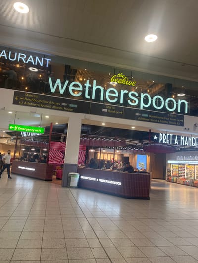The image displays an indoor commercial concourse, likely within a transportation hub given the directional signage and the provided location context of Horley, United Kingdom (near Gatwick Airport). The scene features two prominent food and beverage establishments: "the beehive wetherspoon" and "PRET A MANGER."
The Wetherspoon pub/restaurant, identified by its full name "the beehive wetherspoon" and a partial "AURANT" from "RESTAURANT," occupies the left side of the frame. Nearby, directional signs indicate "Stairs to: Northbound local buses, Ashdown House & Atlantic House" and "Lifts to: Hotel," with similar text repeated on another sign. On the right, "PRET A MANGER" is visible, advertising "ORGANIC COFFEE FRESHLY MADE FOOD." Partially visible text near Pret A Manger includes "Hello London Ba" and "Proudly using freshly made food &." A green "Emergency exit" sign is also present.
Numerous people are visible throughout the image; some are seated inside the establishments, others appear to be working behind counters, and at least one person is walking in the foreground on the tiled floor. The area is brightly lit by artificial overhead lighting on a high ceiling, and the flooring consists of light-colored tiles. The overall activity suggests a busy, functional public space where people are either dining, waiting, or in transit. No specific emotions are discernible, and the time of day is not evident from the indoor setting.
