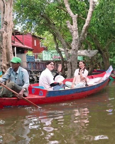In a long, red and blue boat on a body of water, a man in a light blue shirt and green cap rows. Beside him, a person in a white shirt and glasses smiles and waves. In the back of the boat, a young woman in a peach-colored shirt with braided hair also smiles. They are surrounded by lush green trees and vegetation, with a red building visible in the background. The water is calm and reflective, with ripples from the rowing action. This scene appears to be in a floating village in Soutr Nikom, Cambodia, a region known for its stilted houses and waterways.
