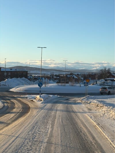 The image depicts a snow-covered roundabout in Kiruna, Sweden, under a clear, light blue sky. The scene is illuminated by low-angle sunlight, casting long shadows.
In the foreground, a snow-covered road with visible tire tracks leads towards the roundabout. Piles of plowed snow line both sides of the road.
The midground features a circular, snow-covered central island. A tall streetlight with twin luminaires stands behind this island. Two blue square signs depicting a white pedestrian symbol above a downward arrow, and a blue circular sign with a white arrow pointing right (indicating mandatory pass-right), are present near the island. To the right, another blue square pedestrian sign and a yellow triangular "Give Way" sign are visible. A light-colored Volvo station wagon, possibly white or silver, is positioned on the right side of the roundabout.
In the background, snow-dusted residential buildings with dark roofs are visible. Beyond these structures, a vast expanse of snow-covered hills and mountains extends to the horizon. At least three wind turbines are identifiable on a distant, snow-covered hill. The sky is clear blue with some horizontally elongated white clouds above the mountain range. No individuals are present in the scene.
