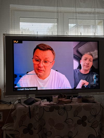 This media file displays a television screen showing a video call or recorded broadcast. Two men are visible. The top portion of the screen shows one man with short, blonde hair and a dark t-shirt, looking towards the camera with a slight smile. The bottom portion shows another man wearing glasses and a white t-shirt, with his head tilted to the side, looking down and slightly to the right. The background of the bottom portion appears to be a plain, light-colored wall with some digital artifacts affecting the image quality. There is text at the bottom right of the screen that reads "олай Осьиченко".
The television is positioned in a room with a window on the left side, covered by sheer white curtains. To the right of the television, a patterned fabric, possibly a tablecloth or bedspread with floral motifs, is draped over furniture. Several objects are placed on a surface near the television, including what appear to be toiletries, a small pink box, and sunglasses.
The lighting suggests it could be daytime, but the room's interior lighting is also a factor. There are no clear indicators of weather or specific time of day beyond general indoor conditions. The activity appears to be a passive viewing experience, with the subjects on the screen engaged in whatever the broadcast or call entails. The emotional tone of the subjects on screen is neutral to slightly positive. The location context of Szczecin, Poland, does not provide specific visual cues within the image itself.