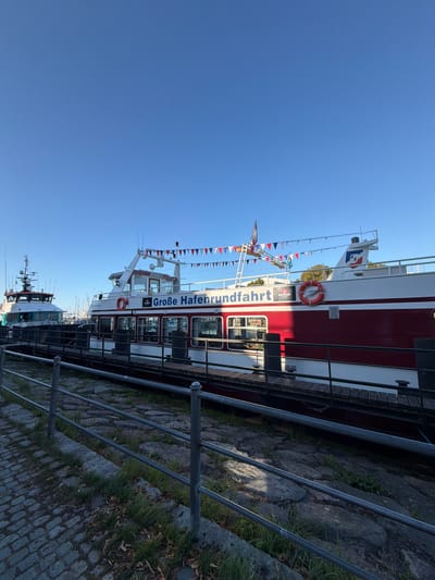 The image shows a bright, clear day with a blue sky. In the foreground, a cobblestone walkway lines a pier with a metal railing. Beyond the railing, two boats are docked. The boat on the right is larger and red and white, with "Große Hafenrundfahrt" (Great Harbor Tour) written on its side in large blue letters. It also has a life preserver and small triangular flags strung along its upper deck. The boat on the left is narrower and appears to be a working vessel with a white superstructure and green hull. In the background, more boats and masts are visible, suggesting this is a harbor or marina. The lighting indicates it is likely morning or late afternoon. The scene appears to be the Warnow River harbor in Rostock, Germany, specifically where harbor tours depart. There are no people visible in the image.