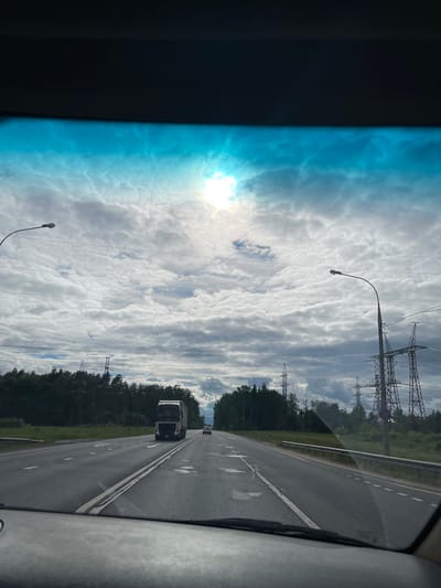 The image depicts a view from inside a moving vehicle on a highway near Nazarovo, Russia. The road has multiple lanes with white dashed lines marking lane divisions. A large truck is visible in the adjacent lane. Overhead power lines and transmission towers run parallel to the road. Trees border the highway. The sky is overcast with clouds. A street light is visible on the left. The sun is partially obscured by the clouds. The foreground shows part of the vehicle's interior, including a blue-tinted window.