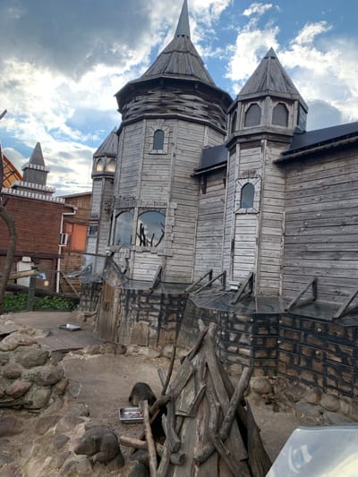 The image depicts a rustic, castle-like structure made primarily of weathered wood, situated in an outdoor enclosure. The structure features multiple turrets with pointed roofs, reminiscent of medieval architecture, and numerous windows of various shapes and sizes. The lower part of the structure is clad in what appears to be dark brick or stone, with wooden supports extending outwards. In the foreground, the ground is a mixture of dirt, rocks, and sand. Several animals, possibly meerkats or similar small mammals, are visible foraging in metal trays. One animal is seen partially obscured by wooden debris in the lower right of the frame. The overall environment suggests an animal exhibit or a themed attraction, possibly within a zoo or park setting. The sky above is cloudy with patches of blue. The location is identified as Малахавецкі сельскі Савет, Belarus.