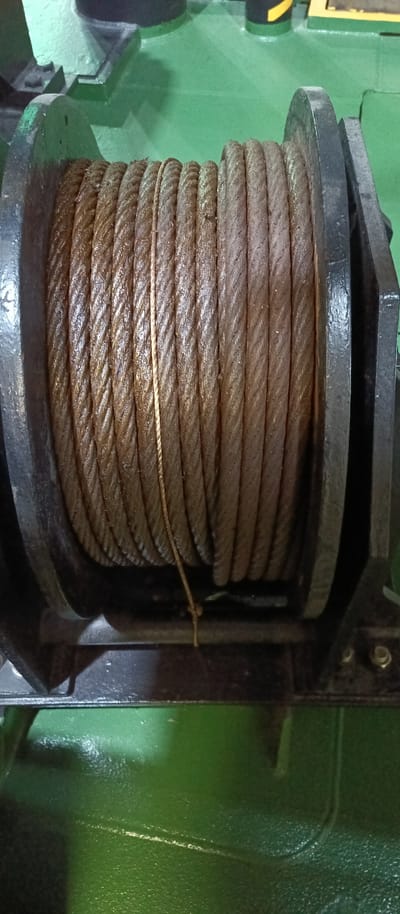 A coiled metal cable is wound around a dark metal spool. The cable appears to be made of multiple strands twisted together and shows signs of wear and some lubrication. The spool is mounted on a black metal frame, which is positioned on a green surface that appears to be a deck. The background is out of focus but suggests an industrial or marine environment, with hints of green and metallic structures. The scene is well-lit, highlighting the texture and details of the cable and spool. There are no people or animals visible in the image. The location in Al Hamriyah, United Arab Emirates, suggests a maritime or industrial setting, possibly a port or shipyard.