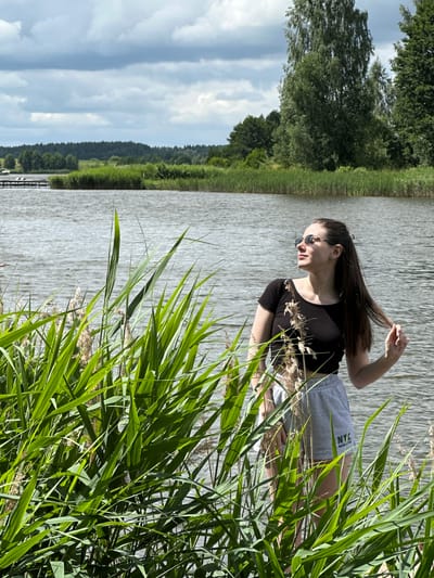 A young woman with long, dark hair and sunglasses stands by a body of water, partially obscured by tall green reeds. She is wearing a black crop top and grey shorts with "NYC MANHATTAN" printed on them. Her hair is pulled back, and she is touching a strand of it with her right hand. The water appears choppy, with trees and a dense treeline visible in the background. The sky is partly cloudy. The location is Вішоўскі сельскі Савет, Belarus.