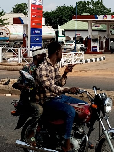 This media file shows two men riding a motorcycle on a street in Garko, Nigeria. The man in front is wearing a checkered shirt and blue jeans, and is pointing with his right hand. The man behind him is wearing a different checkered shirt and a white and black striped cap. They are riding past a gas station with several pumps and a sign displaying fuel prices: PMS, AGO, LPG, and CNG. The gas station appears to be operating, with a white car and a truck visible in the background. The time of day appears to be daytime, with overcast skies. The overall atmosphere is casual, with the individuals focused on their journey. Text visible in the image includes brand names like "DANGOTE A ARANO," "AARANO Lubricant," and "BOXER" on the motorcycle. The price list at the gas station also contains text indicating different fuel types.