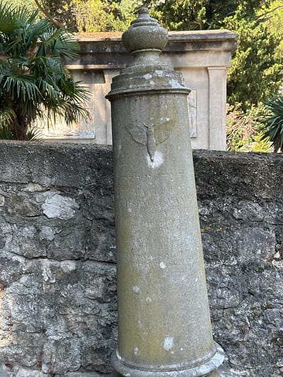 In the city of Škaljari, Montenegro, a weathered, conical stone monument stands tilted to the right, its surface covered in lichen and bearing inscriptions. Atop the monument is a finial, and near its middle, a relief carving of a moth is visible. The monument is positioned in front of a rough, grey stone wall. Behind the wall, a structured building with a sculpted relief is partially visible, flanked by lush green trees and palm fronds. The lighting suggests a sunny day. The inscription on the monument, in a language that appears to be Hebrew and Italian, is partially legible, mentioning dates such as August 1829 and July 1875.