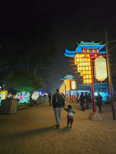 The image depicts an outdoor scene at night, likely in Puente Alto, Chile, characterized by illuminated Asian-style lanterns. The lanterns are multi-tiered, with vibrant colors like blue, red, and yellow. They are hanging above a dirt path with people walking. A person with a backpack is standing on the path. There are trees and other illuminated displays in the background. The scene appears to be part of a festival or cultural event.