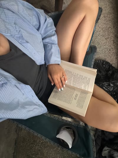 This media file features a close-up, high-angle shot of a person sitting in a camping chair, engrossed in a book. The individual is wearing a light blue striped button-down shirt over a dark grey ribbed top and shorts. Their legs are crossed, with the left leg resting over the right. A book, open to pages filled with text, is held in their hands. The right hand, adorned with a ring and manicured nails painted in pastel colors, is turned towards the camera.
The setting appears to be outdoors, with a speckled, concrete-like ground visible. A dark plastic bag is on the left side of the frame, and the green camping chair has a patterned trim. A cup holder on the chair contains a crumpled piece of white paper.
The text in the book is primarily in Bulgarian. Notable phrases include "Десета глава" (Tenth chapter) and "БЕЛИЯТ ГОСТ" (The White Guest). There are also dates visible, such as "8 януари 2018 г." (January 8, 2018) and "24.11.2009 г." (November 24, 2009).
While no specific time of day or weather conditions are explicitly stated, the presence of a book suggests a leisure activity, possibly during daylight hours. The overall mood conveyed is one of quiet concentration and relaxation.