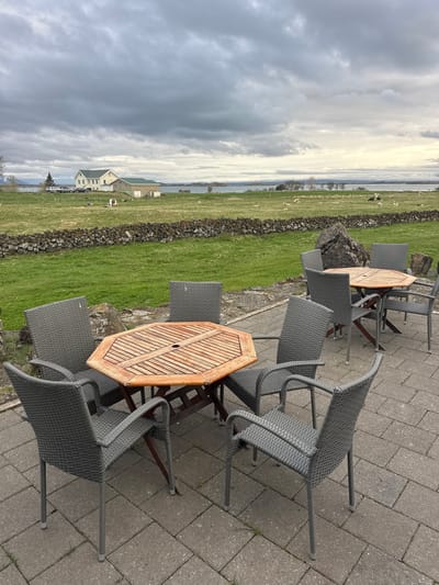 The image displays an outdoor patio with furniture, offering a scenic view of a rural landscape in Reykjahlíð, Iceland. In the foreground, a paved area with grey interlocking stones features several unoccupied hexagonal wooden tables with slatted tops and grey woven-wicker chairs. Some chairs are pushed in, while others are slightly pulled out.

Beyond the patio, a low stone wall, constructed from dark, rough rocks, separates the seating area from a lush green field. Numerous sheep, predominantly white with at least one dark-colored, are scattered across the field, grazing peacefully. In the mid-ground, a calm body of water, likely Lake Mývatn, extends across the frame, reflecting the cloudy sky.

On the far side of the water, several distant buildings with light-colored walls and dark or green roofs are visible, along with parked vehicles, suggesting human habitation. The sky above is largely overcast with patches of brighter clouds, indicating a diffused daylight, possibly late morning or afternoon. The scene evokes a tranquil and inviting atmosphere, showcasing the natural beauty of the Icelandic landscape. No people are present, and there is no visible text.