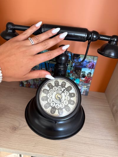 The image shows a black antique rotary telephone indoors. The telephone sits on a wooden surface. In the background, there's an orange wall and a framed picture. In the foreground, a person's hand with white manicured nails and rings is touching the telephone. The rotary dial features both numbers and letters, and it includes the word "TELEPH" and the year "1957." The dial also mentions "DUBLIN." The image provides evidence suggesting a location in Dublin, as noted on the telephone, and possibly a residential or commercial interior setting, but it is not in Les Arcs, France.