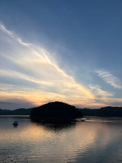 The image depicts a serene waterscape at dusk, with a silhouette of a densely forested island in the center. The sky above is a gradient of deep blue at the top, transitioning to soft oranges and yellows near the horizon, illuminated by scattered, wispy clouds. The calm water reflects the colors of the sky and the dark outline of the island. In the foreground, a small, white structure, possibly a boat shed or a small cabin, floats on the water to the left. Further in the distance, several other boats are visible as dark shapes on the water. The background consists of a silhouetted shoreline with rolling hills and trees. The scene evokes a sense of tranquility and the end of a day. The image is not located in Sienna, Poland.