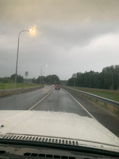 A wide-angle, point-of-view shot from inside a vehicle shows a wet highway under a cloudy sky. The road surface is dark and appears to be wet with rain, with puddles visible. The highway curves to the right, with a metal guardrail on the right side of the road. On the left side of the road, there is a grassy verge and a line of trees. Further down the road, several vehicles are visible, including a red car in the right lane. There are streetlights lining the highway, some of which are illuminated. The sky is overcast with heavy gray clouds, suggesting poor weather conditions. The foreground of the image shows the wet hood and windshield of the vehicle from which the photo was taken, with water droplets visible on the surface. The image appears to have been taken in the city of Neklyudovo, Russia, given the context provided by the prompt, though there are no specific landmarks to confirm this visually.