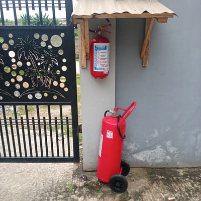 In the city of Ibadan, Nigeria, a scene unfolds in front of a grey concrete wall and a black metal gate with a palm tree motif. Two red fire extinguishers are prominently displayed. One, smaller and cylindrical, hangs from a makeshift wooden shelter attached to the wall. This extinguisher has a white label with blue lettering that reads "POWDER" and instructions for use. The second, larger fire extinguisher stands on two black wheels on the ground, leaning against the wall. It also has a white label with red and blue text, and its hose is coiled around its body. The ground in front of the wall and gate is a mixture of concrete and gravel. In the background, through the gate, vegetation and distant buildings are visible.