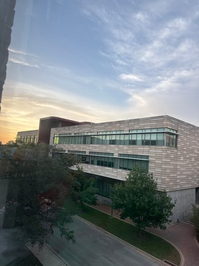 The image depicts a modern building with a beige brick facade and large windows, likely an office or academic building. The building is situated along a street with trees and landscaping. The sky shows a sunset or sunrise, with clouds illuminated by the warm light. The location is likely in Austin, Texas, based on architectural styles and the overall environment. The scene is captured from an elevated position, possibly from another building or a high floor, providing a view over the landscape.