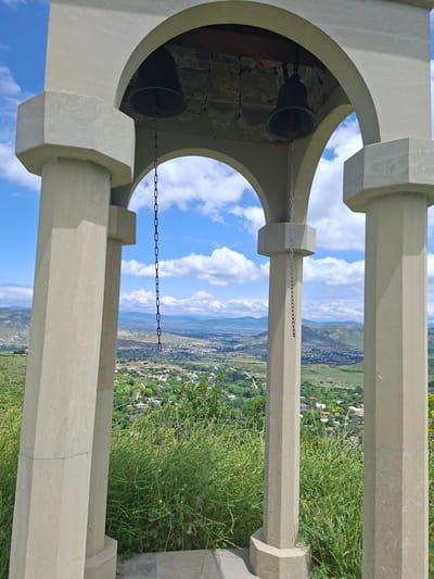 An outdoor, eye-level, medium shot shows a bell tower with a view of the city of Kvemo Nichbisi, Georgia, in the background. The bell tower has two dark bells, each hanging from a metal chain. The tower has four polygonal pillars made of light beige stone, and arched openings. The sky is blue with white clouds. The background is a green landscape with buildings scattered throughout.