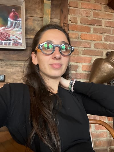 The image displays a head-and-shoulders portrait of a female subject, positioned slightly right of center, looking directly at the camera. She has long, dark brown hair draped over her left shoulder and chest. She wears unique dark-framed, round eyeglasses with reflective lenses, and a black long-sleeved top. Her left wrist, adorned with multiple beaded bracelets including pink and green stones, is visible. Her right hand is placed on the back of her neck.
The background depicts a rustic indoor environment with mixed textures. To the upper left, a framed still-life painting of pomegranates on a clear dish hangs on a dark wooden plank wall. Below this, a black electrical outlet is visible on the wooden wall. To the right of the subject, a red brick wall is present, against which a partial view of a large, bronze-toned vessel with a handle is seen. A vertical wooden beam separates the wooden wall from the brick wall. A curved wooden chair back is partially visible in the lower right foreground. The precise location in the city of Argveta, Georgia, is not verifiable from the visual information provided within the image.