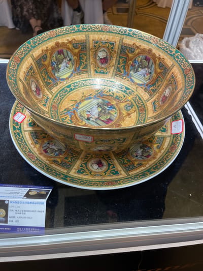 This media file shows a close-up of an ornate porcelain bowl and saucer set, likely displayed in a market or auction setting, possibly in Futian District, China. The porcelain is richly decorated with a gold and teal patterned border featuring vignettes of classical scenes with figures, along with floral motifs. The bowl is placed partially inside the saucer, allowing for a clear view of both items' intricate designs.
Several price tags are visible on the items. One tag, in red and white, appears to have handwritten characters and the number "795". Another red and white tag shows "599". A more detailed white label with blue text, oriented vertically, is in the lower right corner. This label contains information in Chinese and English, identifying the item as part of the "2026 Hong Kong Royal Royal Collection Art Auction". It lists "LOT: 1216" and the title "European Royal Golden Western Figure Story Patterned Bowl and Dish Set". The estimated starting bid is "6,800,000 HKD" and the inscription is "Qing Dynasty". The auction house is identified as "HK Royal Auction Limited".
The background is dark, likely a display surface, with some out-of-focus elements of the surrounding environment, including shelving and perhaps other merchandise. The lighting is focused on the porcelain, highlighting its elaborate decoration and metallic accents. The overall impression is of a high-value, antique art object being presented for sale.