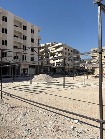 This image depicts an outdoor scene in Mu'adammiyat ash-Sham, Syria, under a clear blue sky. The foreground consists of an unpaved, light-brown dirt and gravel ground with scattered debris, including plastic fragments. Partially erected dark metal frameworks, consistent with market stalls or canopy supports, are prominent, with numerous horizontal metal beams lying on the ground.
In the mid-ground, an unpaved road runs horizontally, separating the open area from the background buildings. A large, conical pile of light-colored aggregate material (gravel or crushed stone) is centrally located. Multiple vehicles are parked along the road. At least three individuals are visible: one walking left-to-right near the aggregate pile, another standing further right on the road, and a third partially visible on the far left.
The background is dominated by multi-story residential or commercial buildings. On the left, a cream-colored building with multiple floors features numerous dark, unfinished window openings and balconies. To its right, another similar building exhibits a more irregular facade with curved balconies and numerous window apertures. Utility poles and power lines are visible between the structures. Strong sunlight from the upper right casts long, distinct shadows of the metal frameworks and individuals onto the ground.