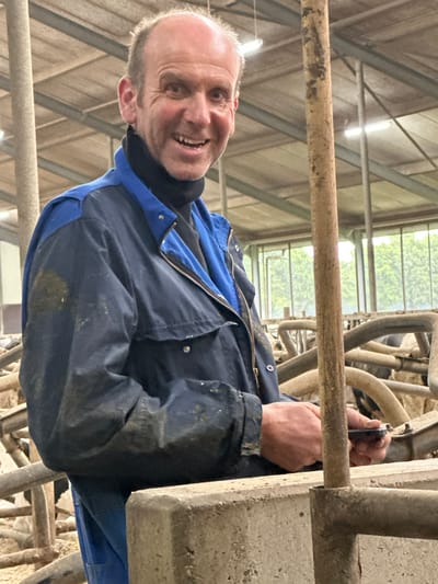 An adult Caucasian male with a shaved head, fair skin, and a broad smile is standing in what appears to be a barn or agricultural setting. He is wearing a dark blue and royal blue work jacket over a black turtleneck. The jacket shows signs of wear and dirt. His right hand is holding a small, dark object, possibly a phone or tool, in front of a concrete partition and metal railings. The background is blurred but suggests an indoor agricultural environment with wooden beams, artificial lighting, and possibly livestock behind the railings. The man's expression is friendly and engaged. The overall impression is of a worker on a farm in Wilhelminaoord, Netherlands, possibly taking a break or interacting with something on his device.