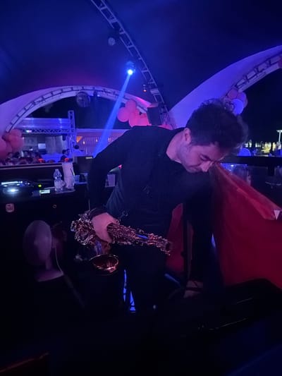 A man stands holding a saxophone in a dimly lit environment. The location appears to be a tent or large structure, as evidenced by the visible supports and lighting fixtures overhead. There are also visible balloons and other decorations suggesting an event or celebration. The exact city of Olgiate Olona, Italy, cannot be confirmed solely from the visual information provided.