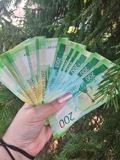 The image shows a hand holding a fan of Russian 200 ruble banknotes, partially obscured by the needles of a fir tree. The background consists of more fir trees and a building. The banknotes are predominantly green and blue, with some featuring a watermark and security thread. The hand has a glittery pink fingernail. The overall lighting suggests it is daytime. The location in Skadovsk, Ukraine, is not discernible from the image itself.