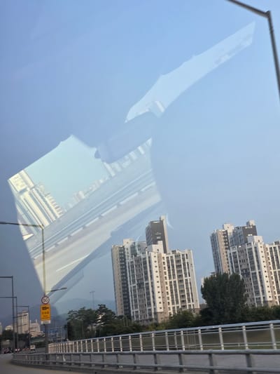 The image captures a view of the city of Daejeon, South Korea, from a moving vehicle. The perspective suggests the photo was taken from a car driving on an elevated road or highway. Dominating the scene are several modern, high-rise apartment buildings, characteristic of urban Korean architecture, with varying facade designs and window patterns. Some buildings have visible text and numbering, such as "VERTUM 202".
A significant element in the foreground is a road barrier with a grey metallic railing, indicative of a bridge or overpass. To the left, there are street lights and traffic signs, including a circular speed limit sign displaying "50" and a rectangular yellow sign with Korean text and "Photo enforcement" below it, warning of speed cameras. A distance marker reading "250m" is also visible on a pole.
The background is filled with a hazy sky, suggesting either early morning, late afternoon, or atmospheric conditions. Faintly visible in the distance are mountains, indicating Daejeon's geographical setting. Reflections on what appears to be a window or windshield are prominent, overlaying the cityscape with distorted images of the sky and possibly the interior of the vehicle or objects outside the frame. Trees are scattered between the buildings and along the roadside, adding a touch of nature to the urban environment. The overall impression is a typical urban commute scene in a South Korean city, with a blend of modern infrastructure and natural elements.