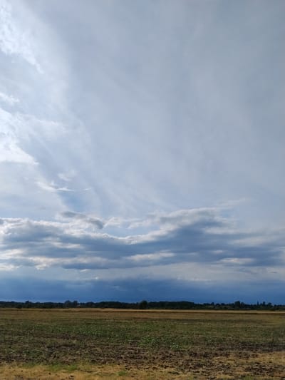 This is a vertical, outdoor, medium-shot photograph taken in Ustyvytsya, Ukraine. The image depicts a rural landscape under a dramatic sky. The foreground shows a plowed field with patches of dry grass and some green vegetation. Beyond this field, a line of dark green trees forms the horizon. Above the trees, the sky is filled with a mix of clouds. The lower portion of the sky features dense, dark clouds that appear heavy and ominous, with hints of sunlight breaking through in some areas. Higher up, the sky transitions to lighter, wispy clouds that are illuminated by sunlight, creating streaks and patterns across the expanse. The overall impression is one of a vast, open space with a dynamic sky.
