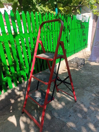 A red, three-step stepladder is situated outdoors on a concrete surface in front of a bright green wooden fence. The fence has vertical planks with pointed tops. Sunlight casts dappled shadows on the ground from the surrounding foliage and the ladder itself. To the right of the ladder, near the fence, is a black plastic crate filled with what appears to be assorted items, possibly fruits or vegetables. In the background, beyond the fence, is vegetation and part of a building. The scene is well-lit, indicating it is daytime. The image does not contain any people or show any actions.
