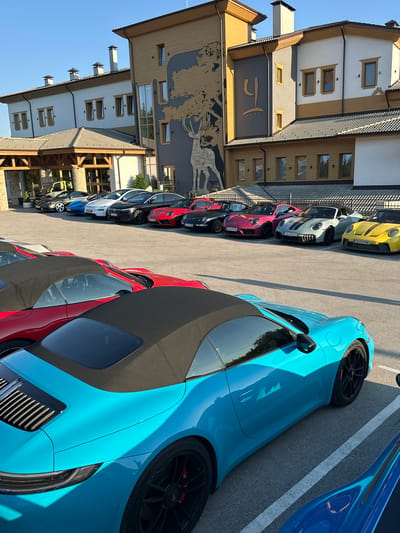 The image depicts a parking lot in front of a building that appears to be a hotel or a similar establishment, located in Kerenite, Bulgaria. The building has a modern architectural design with elements of wood and stone. A large mural of a deer is visible on the facade of the building. A row of high-performance sports cars, predominantly Porsches, are parked in the lot. The cars are of various colors, including turquoise, red, pink, gray, and yellow. The scene is captured on a clear, sunny day. No people are visible in the image. The cars are parked in designated parking spaces. The environment suggests a luxury resort or a gathering of car enthusiasts.